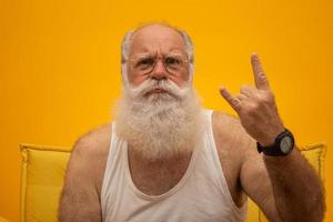 Old man with a long beard on a yellow background. Hand giving the devil horns gesture. Senior with full white beard. Old man with a long beard with sadness. photo