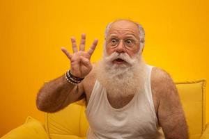 senior sonriente con un hombre de barba blanca larga haciendo cuatro veces el gesto de señas con los dedos de la mano sobre fondo amarillo. emoción positiva expresión facial sentimientos, actitud, símbolo, cuenta regresiva. foto