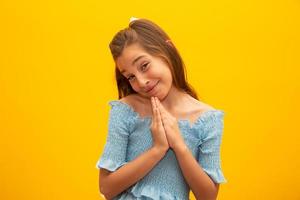 Nice little girl, holds hands in praying gesture, stands over isolated yellow background with copy space. photo