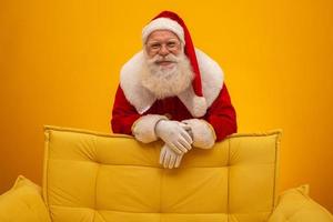 Santa Claus sitting on a yellow couch on yellow background with copy space. Yellow sofa. photo