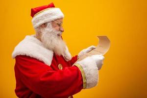 Happy Santa Claus holding vintage paper on yellow background. Merry Christmas and New Year's Eve concept. Copy space. Difficulty reading. Ugly handwriting. photo