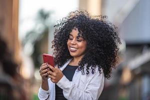 joven mujer negra de pelo rizado caminando con teléfono celular. enviando mensajes de texto en la calle. Gran ciudad. foto