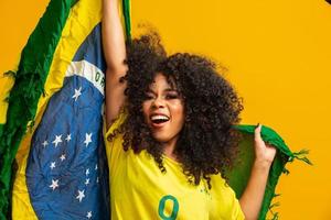 chica afro animando al equipo brasileño favorito, sosteniendo la bandera nacional en fondo amarillo.