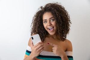 Studio shot of entertained cute happy african american girl with afro hairstyle holding smartphone using device to have fun photo