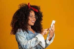 Studio shot of entertained cute happy african american girl with afro hairstyle holding smartphone using device to have fun. Yellow background. photo