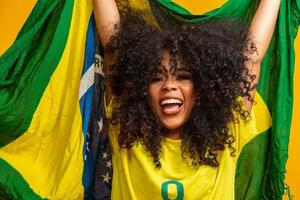 chica afro animando al equipo brasileño favorito, sosteniendo la bandera nacional en fondo amarillo. foto