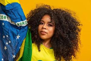 chica afro animando al equipo brasileño favorito, sosteniendo la bandera nacional en fondo amarillo. foto
