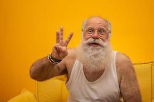 senior sonriente con un hombre de barba blanca larga haciendo tres veces el gesto de firmar con los dedos de la mano sobre fondo amarillo. emoción positiva expresión facial sentimientos, actitud, símbolo, cuenta regresiva. foto