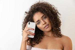 Studio shot of entertained cute happy african american girl with afro hairstyle holding smartphone using device to have fun photo