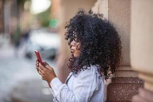 Young curly hair black woman walking using cell phone. Texting on street. Big city. photo