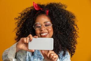 Studio shot of entertained cute happy african american girl with afro hairstyle holding smartphone using device to have fun. Yellow background. photo