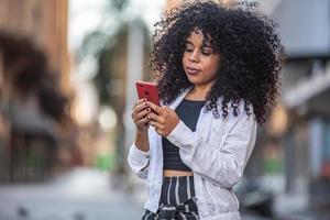 Young curly hair black woman walking using cell phone. Texting on street. Big city.