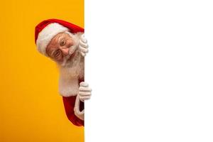 Happy Santa Claus looking out from behind the blank sign isolated on yellow background with copy space photo