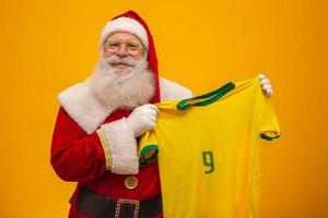 santa claus es fanático de brasil. partidario de santa claus del equipo brasileño. campeonato deportivo santa claus sosteniendo la camiseta del equipo. partido de fútbol. foto