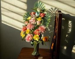 Bouquet of fake artificial roses in a vase set on a wooden chair with louvered sunlight. photo