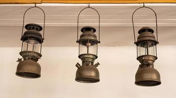 Three old storm lanterns are hung from hooks on a white concrete wall. photo