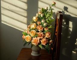 Bouquet of fake artificial roses in a vase set on a wooden chair with louvered sunlight. photo