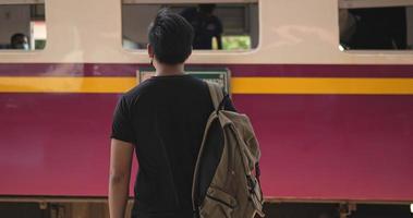 Back view of Young Asian traveler man waiting the train at train station. Male wearing protective masks, during Covid-19 emergency. Transportation, travel and social distancing concept.