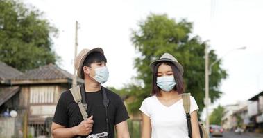 vista frontal do casal feliz viajante asiático com mão de chapéu juntos enquanto caminhava na rua. sorridente jovem falando com mulher. casal usando máscaras de proteção, durante a emergência covid-19.