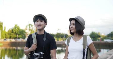 Vorderansicht des glücklichen asiatischen Reisendenpaares mit Huthand zusammen beim Gehen im Park. lächelnder junger Mann und Frau mit Blick auf den Park. urlaubs-, reise- und hobbykonzept. video