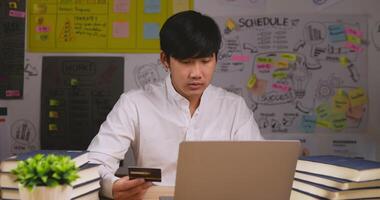 portrait d'un jeune homme asiatique a appuyé sur le code de la carte de crédit pour payer en ligne via un ordinateur portable au bureau dans la chambre. homme tenant une carte de crédit et faisant des achats en ligne. achats en ligne et concept de commerce électronique. video