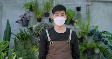 Portrait of Asian male gardener owner in face mask is crossing arm and looking at camera in the garden. Home greenery, selling online and hobby concept. video
