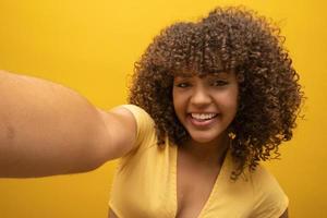 Close up of excited young african american girl in fur sweater posing isolated on yellow orange background in studio. People lifestyle concept. Mock up copy space. Doing selfie shot on mobile phone. photo