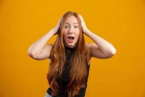 Young redhead woman raising hands to head, open-mouthed, feeling extremely lucky, surprised, excited and happy against yellow wall. photo