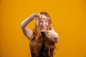 mujer caucásica pelirroja sobre fondo amarillo aislado sonriendo haciendo marco con manos y dedos con cara feliz. concepto de creatividad y fotografía. cineasta o fotógrafo. idea de visión de lente. foto