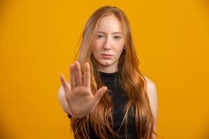 Beautiful redhead woman standing over isolated yellow background with open hand doing stop sign with serious and confident expression, defense gesture. No more violence against women. Abuse. photo