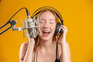 Beautiful redhead woman face singing with a condenser silver microphone open mouth performing song pose over yellow background copy space for your text. Fm radio announcer.