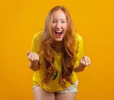 partidario de brasil. hincha brasileña pelirroja celebrando el fútbol, partido de fútbol con fondo amarillo. colores de brasil. foto
