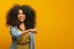 Cheerful Afro woman points away on copy space, discusses amazing promo, gives way or direction, wears yellow warm sweater, has pleasant smile, feels optimistic, isolated over yellow background.