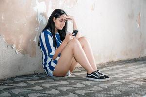 Single sad teen holding a mobile phone lamenting sitting on the sidewalk. Crying depressed teen girl holds phone sitting on the sidewalk. photo