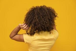 Backwards American African woman with her curly hair on yellow background. Laughing curly woman in sweater touching her hair and looking at the camera.