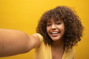 Close up of excited young african american girl in fur sweater posing isolated on yellow orange background in studio. People lifestyle concept. Mock up copy space. Doing selfie shot on mobile phone. photo