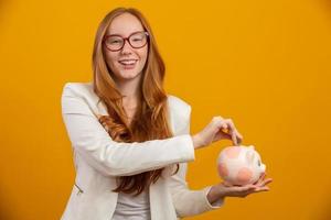 A redhead young woman with a pink pig coin bank, money box, for banking, saving, deposit. Isolated yellow background. photo