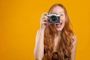 Photo of adorable redhead woman wearing black clothes holding retro vintage camera and taking picture isolated over yellow background. Mock up copy space. Hold retro vintage photo camera.