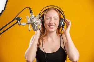 Beautiful redhead woman face singing with a condenser silver microphone open mouth performing song pose over yellow background copy space for your text. Fm radio announcer. photo