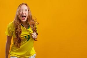partidario de brasil. hincha brasileña pelirroja celebrando el fútbol, partido de fútbol con fondo amarillo. colores de brasil.