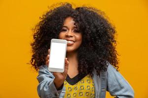 retrato de una joven alegre, positiva y atractiva con pantalones de jeans, con un teléfono inteligente con pantalla blanca en la mano, señalando con el dedo índice al producto, aislada en un fondo amarillo