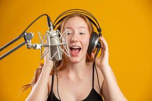 Beautiful redhead woman face singing with a condenser silver microphone open mouth performing song pose over yellow background copy space for your text. Fm radio announcer. photo