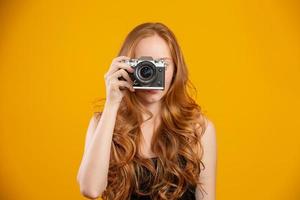 Photo of adorable redhead woman wearing black clothes holding retro vintage camera and taking picture isolated over yellow background. Mock up copy space. Hold retro vintage photo camera.