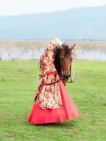 A beautiful western woman in a beautiful dress stands with her horse in the lake meadow photo