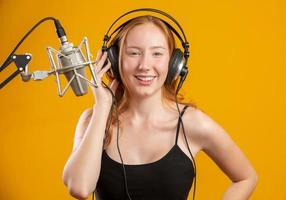 Beautiful redhead woman face singing with a condenser silver microphone open mouth performing song pose over yellow background copy space for your text. Fm radio announcer. photo
