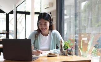 Smiling girl student wear wireless headphone study online with skype teacher, happy young woman learn language listen lecture watch webinar write notes look at laptop sit in cafe, distant education photo
