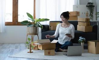 Smiling young Asian business owner woman prepare parcel box and standing check online orders of product for deliver to customer on laptop computer. Shopping Online concept. photo