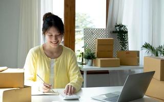 A portrait of Asian woman, e-commerce employee sitting in the office full of packages on the table using a calculator, for SME business, e-commerce, technology and delivery business. photo