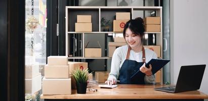 una joven y sonriente propietaria de una empresa asiática prepara una caja de paquetes y revisa de pie los pedidos de productos en línea para entregarlos al cliente en una computadora portátil. concepto de compras en línea. foto