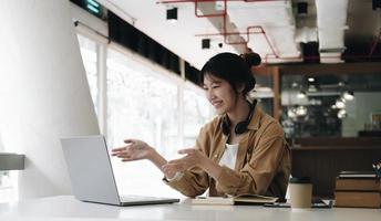 Freelance business women casual wear using laptop working call video conference with customer in workplace in living room at home. Happy young Asian girl relax sitting on desk do job in internet. photo